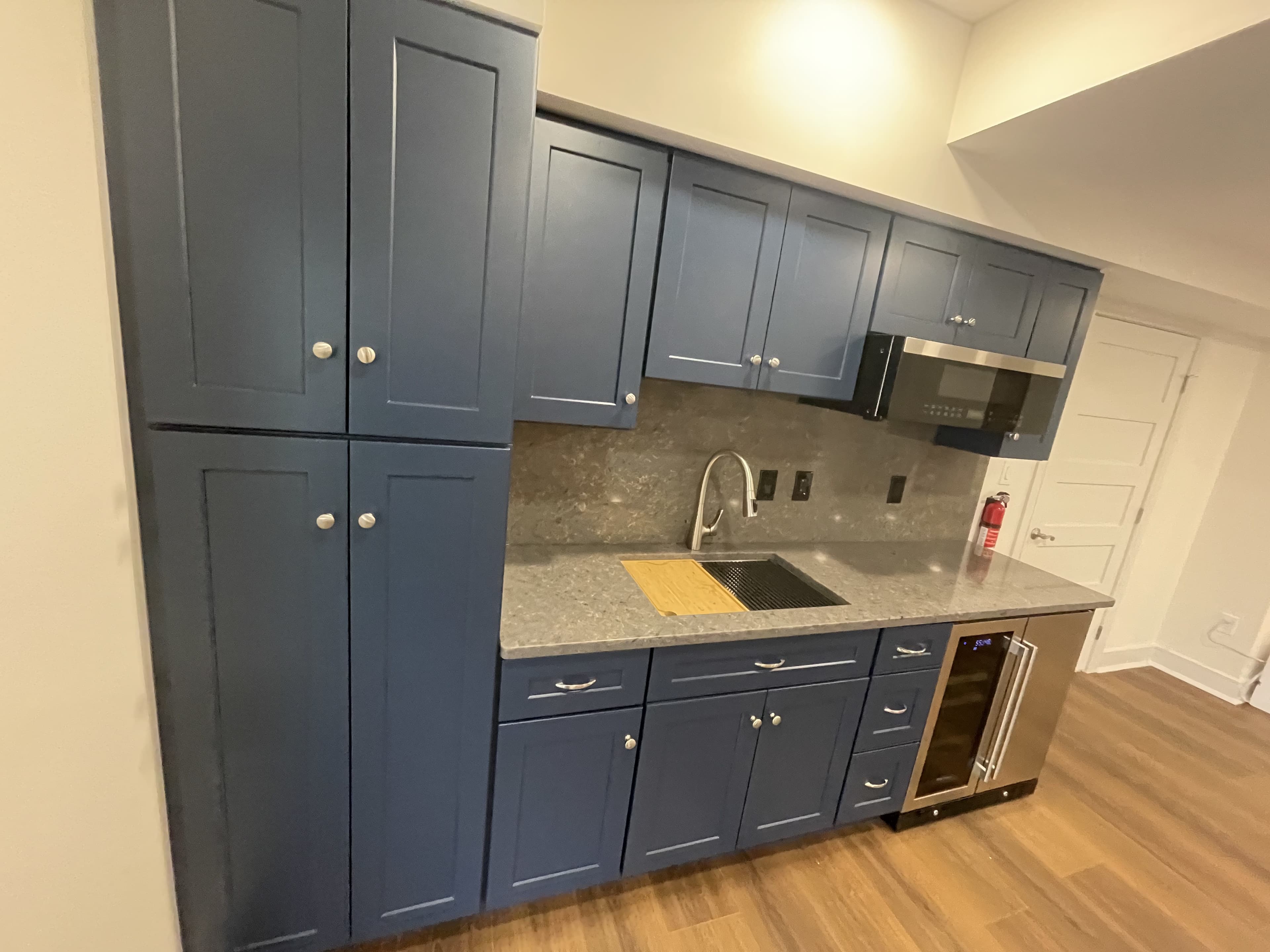 Wide view of blue cabinetry with a gray countertop and backsplash, sink and faucet, microwave and beverage cooler.