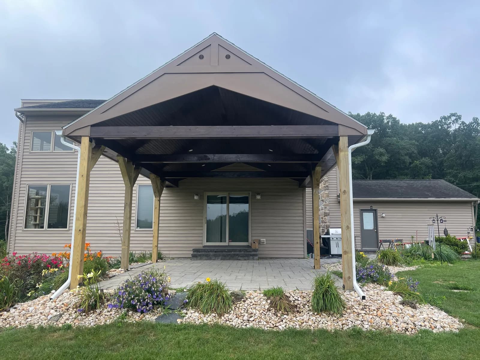 Front view of a gable-roof pavilion, patio, and planted garden edges beside a house.